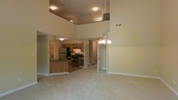 This two-story residence offers an open-concept design that maximizes the sense of space and flow throughout the main living areas. The living room is expansive, featuring impressively tall ceilings that extend upward to reveal a second-floor overlook, adding a dramatic architectural element to the interior. Soft, neutral-toned carpeting covers the main floor, contributing to a warm and inviting ambiance. A chandelier with multiple light fixtures serves as a focal point, providing both style and illumination to the open space. The kitchen is accessible directly from the living area and is outfitted with warm wood-toned cabinetry, stainless steel appliances including a refrigerator and gas range, and dark countertops. A kitchen island provides additional workspace and storage. An interior staircase connects the main floor to the upper level, where a landing overlooks the living space below. The upper floor features its own recessed lighting and railing, reinforcing the home's open vertical design. Neutral wall tones throughout complement the overall layout, creating a cohesive and functional living environment across both floors.