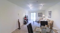 This well-appointed single-floor residence offers a thoughtfully designed open-concept layout that seamlessly connects the dining and living areas, creating a spacious and inviting atmosphere. The interior is bathed in natural light, thanks to a large sliding glass door that leads to a private balcony, offering pleasant outdoor views and a welcoming sense of openness.

The living area features plush, light-toned carpeting underfoot, contributing to the warm and comfortable ambiance throughout the space. A stylish glass coffee table anchors the seating area, which is furnished with a neutral-toned sofa and coordinating armchair, creating an elegant yet relaxed setting. A contemporary open shelving unit provides both display and storage space, showcasing decorative accents that add character to the room.

Overhead, modern track lighting is installed across the ceiling, providing flexible and effective illumination throughout the open-plan area. The crisp white walls enhance the brightness of the space, making it feel airy and expansive.

The dining area is defined by a sleek glass-top table paired with dark, high-backed chairs, offering a refined contrast to the lighter living space. Together, the areas form a cohesive and functional living environment with a clean, contemporary aesthetic.