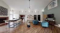 This single-floor clubhouse-style space is designed to serve as a welcoming communal gathering area, combining several functional zones within one expansive, open-concept layout. The architectural highlight of the space is its soaring vaulted ceiling, which lends an airy, grand atmosphere throughout. A striking sputnik-style chandelier hangs prominently above the main lounge area, complementing the contemporary design aesthetic.

The lounge area features plush teal velvet armchairs arranged around a round white accent table, set against a sage green accent wall. A wall-mounted flat-screen television and a wooden media console complete this relaxed seating zone, making it well-suited for casual gatherings. Woven geometric wall art provides a textural focal point along the adjacent white wall.

To one side, a long communal dining bar with counter-height seating accommodates group dining or collaborative work. Natural light filters in through large windows, offering views of surrounding greenery. A dedicated dining nook with a round dark wood table and glass-panel door further extends the dining options.

The fully equipped kitchen features rich cherry wood cabinetry, granite-look countertops, stainless steel appliances including a side-by-side refrigerator and electric range, and tile flooring. Throughout the main areas, durable wood-look flooring ties the entire space together cohesively.