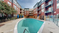 This outdoor pool area in Alexandria, Virginia, is a well-appointed amenity space nestled within a courtyard formed by a multi-story residential building. The architecture surrounding the pool features a striking combination of bold red horizontal siding and contrasting gray paneling, with multiple balconies overlooking the space and large windows that contribute to an open, airy atmosphere.

The generously sized pool itself features clear turquoise water with tiled edges and is accessible via entry steps and handrail access points. The pool deck is finished in smooth concrete and provides ample space for lounging and circulation. Blue market umbrellas are distributed around the perimeter, offering shaded seating options throughout the day.

The seating areas include a mix of lifeguard chairs, benches, and contemporary concrete accent furniture, creating functional rest zones along the pool's edges. Landscaped greenery, including mature trees and planted borders, softens the hardscape and adds natural character to the space. A decorative pergola structure is visible near the building entrance at one end of the pool, marking a focal point within the courtyard. The area is enclosed by an ornamental iron fence with a gated entry, ensuring both safety and privacy.