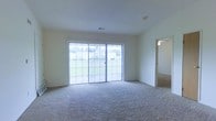 This single-floor space offers a clean, functional layout designed with simplicity and comfort in mind. The main living area is a standout feature, characterized by its generous proportions and wall-to-wall carpeting that adds warmth underfoot. Large sliding glass doors span a significant portion of one wall, bathing the room in abundant natural light and providing a seamless visual connection to the outdoors, where greenery is visible beyond. The neutral color palette throughout — from the soft carpet to the crisp white walls and ceiling — creates a bright, airy atmosphere that feels open and inviting. A ceiling-mounted light fixture is visible in the adjoining room, which is accessible through a wide doorway on the right side of the living area, hinting at a connected and flowing floor plan. Overhead, a centrally placed ceiling vent speaks to a well-integrated climate control system. The overall ambiance of the space is calm and understated, providing a versatile canvas well-suited for a variety of uses and personal styling preferences.