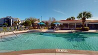 Located along U.S. 98 in Pensacola, Florida, this single-story outdoor amenity complex offers a resort-style experience designed for relaxation and recreation. The centerpiece is a generously sized freeform swimming pool with a smooth curved edge, surrounded by a broad concrete sun deck lined with lounge chairs and blue market umbrellas that provide ample shade throughout the day. The pool area is framed by a Mediterranean-inspired clubhouse building featuring a distinctive red tile roof, large floor-to-ceiling windows, and French doors that open directly onto the poolside patio — allowing natural light to flood both indoor and outdoor spaces seamlessly. Manicured landscaping, including palm trees and trimmed shrubs set against mulched garden beds, adds a tropical character to the environment. A separate, expansive pool with an extended sundeck and additional rows of lounge seating provides further space for recreation, backed by a natural tree line that offers privacy and greenery. A covered gazebo pavilion with ceiling fans, bar-height seating, and column detailing serves as a social gathering point within the complex. The grounds also include a playground area and open grassy spaces, completing a well-rounded outdoor amenity hub suited for community use in the Florida sunshine.