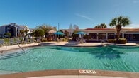 Located along U.S. 98 in Pensacola, Florida, this single-story outdoor amenity complex offers a resort-style experience designed for relaxation and recreation. The centerpiece is a generously sized freeform swimming pool with a smooth curved edge, surrounded by a broad concrete sun deck lined with lounge chairs and blue market umbrellas that provide ample shade throughout the day. The pool area is framed by a Mediterranean-inspired clubhouse building featuring a distinctive red tile roof, large floor-to-ceiling windows, and French doors that open directly onto the poolside patio — allowing natural light to flood both indoor and outdoor spaces seamlessly. Manicured landscaping, including palm trees and trimmed shrubs set against mulched garden beds, adds a tropical character to the environment. A separate, expansive pool with an extended sundeck and additional rows of lounge seating provides further space for recreation, backed by a natural tree line that offers privacy and greenery. A covered gazebo pavilion with ceiling fans, bar-height seating, and column detailing serves as a social gathering point within the complex. The grounds also include a playground area and open grassy spaces, completing a well-rounded outdoor amenity hub suited for community use in the Florida sunshine.