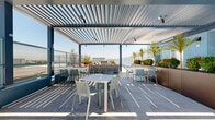 Located atop the building at 1642 E Florence Ave in Los Angeles, this rooftop terrace offers a well-appointed outdoor amenity space with sweeping panoramic views of the surrounding cityscape under Southern California's open blue skies. The terrace is divided into distinct functional zones, each designed for comfort and social engagement.

The open-air lounge area features modern, rust-toned armchairs paired with a sleek blue sofa and a round white side table, arranged to encourage conversation while taking in the expansive views. Large Corten steel planters filled with ornamental shrubs and slender trees are positioned throughout, adding greenery and a naturalistic design element to the rooftop environment.

A covered pergola section provides a shaded dining area, where light filters dramatically through the louvered roof structure, casting geometric shadow patterns across the concrete deck. Light-toned dining tables and chairs furnish this area, making it suitable for both casual gatherings and structured seating.

The terrace is finished with large-format, light-colored porcelain tiles throughout, lending a clean and cohesive aesthetic. Teal-toned mesh perimeter fencing ensures safety while preserving the open sightlines. HVAC equipment is neatly enclosed within designated screened sections, maintaining the overall visual appeal of the space.