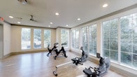 This single-floor fitness space is thoughtfully designed to inspire an active lifestyle. The room is bathed in natural light, courtesy of expansive floor-to-ceiling windows that line multiple walls, offering serene views of lush, tree-filled surroundings. The abundance of glazing creates an open, airy atmosphere that makes the space feel connected to the outdoors while you exercise. Smooth, light-toned hardwood flooring runs throughout, providing both a clean aesthetic and a practical surface for a variety of workout routines. Recessed ceiling lighting supplements the natural light, ensuring the space remains well-illuminated at any time of day. A ceiling fan adds comfort by promoting air circulation during intense workouts. The room accommodates a range of cardio equipment, including stationary cycling bikes and a rowing machine, making it well-suited for diverse fitness regimens. The neutral wall tones and clean architectural lines contribute to a calm, distraction-free environment. Overall, this fitness space strikes a balance between functional design and a bright, welcoming ambiance — an ideal setting for daily exercise and wellness.