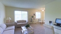 This single-floor space offers a comfortable and well-lit living environment designed for easy, everyday living. The main living area is generously sized, featuring plush wall-to-wall carpeting that adds warmth underfoot. Large windows line the walls, flooding the room with natural light and offering pleasant outdoor views of the surrounding greenery, creating an airy and open atmosphere throughout.

The living area is thoughtfully laid out, with ample room for seating and relaxed gatherings. A ceiling fan is centrally positioned, providing both functional air circulation and a subtle decorative touch. The open-arch display shelving unit serves as a tasteful divider and focal point, adding a decorative element to the space.

Adjacent to the living area, a dining space is visible, accommodating a glass-top table and chairs, ideal for casual meals or entertaining. The light-toned walls throughout contribute to the overall brightness of the interior, complementing the natural light streaming in from multiple windows. The textured ceiling adds a classic architectural detail. Overall, this space combines practicality with a warm, inviting ambiance, making it a comfortable and functional single-floor residence.