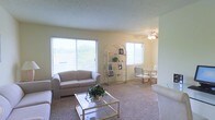 This single-floor space offers a comfortable and well-lit living environment designed for easy, everyday living. The main living area is generously sized, featuring plush wall-to-wall carpeting that adds warmth underfoot. Large windows line the walls, flooding the room with natural light and offering pleasant outdoor views of the surrounding greenery, creating an airy and open atmosphere throughout.

The living area is thoughtfully laid out, with ample room for seating and relaxed gatherings. A ceiling fan is centrally positioned, providing both functional air circulation and a subtle decorative touch. The open-arch display shelving unit serves as a tasteful divider and focal point, adding a decorative element to the space.

Adjacent to the living area, a dining space is visible, accommodating a glass-top table and chairs, ideal for casual meals or entertaining. The light-toned walls throughout contribute to the overall brightness of the interior, complementing the natural light streaming in from multiple windows. The textured ceiling adds a classic architectural detail. Overall, this space combines practicality with a warm, inviting ambiance, making it a comfortable and functional single-floor residence.