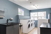 Located at 75 Stewart St in Oakville, Ontario, this shared laundry facility is a compact, single-floor space designed for practical, everyday use. The main laundry room is finished with calming muted blue-grey walls, light wood-look flooring, and fluorescent ceiling lighting that ensures a bright and functional environment throughout.

The main area is lined on both sides with rows of white front-loading washers and dryers, providing ample capacity for resident use. Along one wall, a dark granite-topped counter with built-in cabinetry offers convenient folding and sorting space. A deep utility sink with a modern matte black faucet is integrated into the countertop, accompanied by a wall-mounted paper towel dispenser and soap dispenser for added convenience. A wall-mounted payment terminal and posted instructions make operating the machines straightforward.

A window at the far end of the main room allows natural light to filter in, offering a pleasant view of surrounding trees. An adjoining room provides access to a washroom, adding to the overall functionality of the space. The layout is thoughtfully organized to allow easy movement between machines and work surfaces, making this laundry facility a clean, efficient, and user-friendly amenity.
