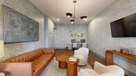 Located at 2555 Cobb Pkwy in Kennesaw, Georgia, this thoughtfully designed single-floor community lounge seamlessly blends comfort, collaboration, and productivity into one cohesive space. The room is unified by striking soft-blue botanical wallcoverings with a subtle metallic sheen, lending an organic yet sophisticated ambiance throughout.

The lounge area anchors the space with a tufted cognac leather sofa, plush cream boucle accent chairs, and a pair of cylindrical wooden coffee tables arranged atop a neutral area rug. An abstract canvas artwork and a wall-mounted flat-screen television add visual interest, while a sculptural multi-arm chandelier provides warm, focused overhead lighting.

Adjacent to the lounge, a dedicated conference nook features a rectangular wood table surrounded by rolling office chairs, bathed in natural light from a large picture window dressed with floor-length drapes. Two sleek black pendant lights hang above, reinforcing the modern aesthetic.

A built-in business center lines one wall, offering dual computer workstations set against the botanical wallcovering, accented by a series of gold sculptural wall pieces above. Rich wood-look flooring flows seamlessly throughout the entire space, grounding each functional zone while maintaining a warm, inviting character. This versatile room is well-suited for socializing, focused work, or small group meetings.