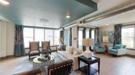 This business center is a thoughtfully designed single-floor space that seamlessly blends professional functionality with contemporary style. The lounge area serves as the centerpiece, featuring a distinctive teal-painted textured ceiling with exposed silver ductwork that lends an industrial-modern character to the space. Large windows line the perimeter, flooding the interior with abundant natural light and offering views of the surrounding urban environment. Patterned wingback chairs in teal and tan tones are paired with a plush cream sofa, arranged around a circular gold accent table atop a geometric area rug, creating an inviting setting for meetings or relaxed collaboration. Light wood-toned flooring and dark-framed glass doors contribute to the space's polished aesthetic. A wall-mounted flat-screen television is integrated into a wood accent wall, adding a media-ready element to the lounge. The kitchenette is well-appointed with dark cabinetry, white quartz countertops, stainless steel appliances including a refrigerator, built-in ovens, and a premium coffee station. Workstations with computer setups are positioned near natural light, providing a dedicated area for focused tasks. The overall ambiance is modern, energetic, and professionally equipped.