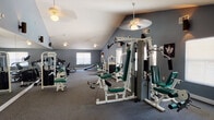 This single-floor community amenity space is thoughtfully divided into two functional areas: a generously sized fitness center and a dedicated laundry facility. The fitness center is the standout feature, characterized by dramatic vaulted ceilings that create an open, airy atmosphere. The walls are finished in a cool slate-blue tone and lined with large mirrors that amplify the sense of space. The room is outfitted with a comprehensive selection of equipment, including multiple treadmills, stationary and recumbent bikes, a multi-station cable machine, a lat pulldown machine, a mini trampoline, and free weight areas. Ceiling fans are distributed throughout to maintain comfortable airflow during workouts. Natural light filters in through several windows, and a glass-panel door provides direct access to an outdoor pool area, offering a refreshing view of the surrounding landscape. The floor is covered in durable grey carpet suited for fitness use. The adjacent laundry facility features a bright, clean environment with multiple stacked washer and dryer units, vinyl flooring, and fluorescent lighting, providing a practical and efficient space for residents' everyday needs.