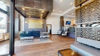 The lobby and lounge at Rowe Residences in Victoria, MN is a generously scaled, single-floor common area that makes a strong architectural statement from the moment you enter. The reception area greets visitors with a dramatic coffered tin ceiling accented by pendant brass lighting, a sleek marble-faced reception desk, and large floor-to-ceiling windows that flood the space with natural light and offer views of the surrounding streetscape. Warm hardwood flooring runs throughout, tying together the many distinct zones within the lounge.

The main lounge area is anchored by a striking floor-to-ceiling black tile fireplace wall with an integrated flat-screen display, surrounded by curated seating arrangements featuring curved accent chairs and a navy sectional sofa. A separate, more intimate lounge nook is appointed with wainscoted walls, rich patterned carpeting, a tufted pink velvet sofa, and framed black-and-white artwork, evoking a boutique hotel aesthetic.

Additional gathering areas include an open dining and community room with tray ceilings, arched architectural details, and a credenza, as well as a bright sitting room with soaring double-height windows. A kitchen and staircase complete the functional layout, making this a thoughtfully designed, community-centered space.
