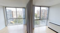 This single-floor space is defined by its abundance of natural light, courtesy of large, wraparound windows that frame dynamic urban views. The windows overlook a cityscape of mid-rise brick buildings interspersed with mature trees, offering a scenic and lively backdrop. Floor-to-ceiling curtains in a soft neutral tone are mounted along a ceiling track, providing flexibility to control light and privacy throughout the space. The smooth hardwood flooring adds warmth and continuity to the interior, complementing the clean, minimalist aesthetic. The overall ambiance is airy and open, with the generous glazing creating a seamless connection between the interior and the surrounding urban environment. The neutral palette of the curtains and flooring allows for versatile styling, making the space highly adaptable. The elevated vantage point further enhances the sense of openness, with treetop and rooftop views contributing to a bright, elevated living experience. The space presents a functional and light-filled environment well-suited to a variety of uses.