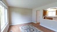 This multi-floor space spans three levels and presents a clean, functional layout with a neutral aesthetic throughout. The living area features warm hardwood-style flooring paired with soft, greige-toned walls and crisp white baseboards, creating a bright and versatile backdrop suitable for a variety of furnishing styles. A doorway leads to additional rooms, and an open pass-through connects the living space to the kitchen, enhancing the sense of flow between areas.

The kitchen is well-appointed with warm honey-toned wood cabinetry offering generous storage above and below the counters. Stone-look countertops provide a durable and attractive work surface, complemented by a double stainless steel sink positioned beneath a large window. This window allows ample natural light to fill the kitchen while providing a view of the surrounding outdoor greenery and neighborhood. A dishwasher is integrated into the cabinetry, adding convenience to the space.

Overall, the space maintains a cohesive, understated design with practical finishes throughout. The combination of natural light, warm wood tones, and neutral walls contributes to a welcoming and livable ambiance across all three floors.