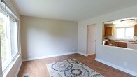 This multi-floor space spans three levels and presents a clean, functional layout with a neutral aesthetic throughout. The living area features warm hardwood-style flooring paired with soft, greige-toned walls and crisp white baseboards, creating a bright and versatile backdrop suitable for a variety of furnishing styles. A doorway leads to additional rooms, and an open pass-through connects the living space to the kitchen, enhancing the sense of flow between areas.

The kitchen is well-appointed with warm honey-toned wood cabinetry offering generous storage above and below the counters. Stone-look countertops provide a durable and attractive work surface, complemented by a double stainless steel sink positioned beneath a large window. This window allows ample natural light to fill the kitchen while providing a view of the surrounding outdoor greenery and neighborhood. A dishwasher is integrated into the cabinetry, adding convenience to the space.

Overall, the space maintains a cohesive, understated design with practical finishes throughout. The combination of natural light, warm wood tones, and neutral walls contributes to a welcoming and livable ambiance across all three floors.