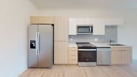 Located at 1824 N Brevard St in Charlotte, NC, this single-floor unit showcases a clean, contemporary design throughout. The open-concept living and kitchen area is bathed in natural light from a large floor-to-ceiling window that frames outdoor views of the surrounding neighborhood, creating a bright and airy atmosphere.

The kitchen is thoughtfully designed with a combination of white upper cabinetry and warm light wood lower cabinets, complemented by a soft gray subway tile backsplash. A white countertop houses a double-basin stainless steel sink, while a full suite of stainless steel appliances — including a side-by-side refrigerator, range, over-the-range microwave, and dishwasher — provides a polished, modern look.

Light wood-tone luxury vinyl plank flooring runs consistently throughout the unit, adding warmth and visual continuity to every room. The bedroom is a calm, well-proportioned space featuring a natural wood door and a window that brings in additional natural light.

The bathroom is sleek and functional, offering a floating white vanity with an extended countertop, a large mirror, a glass-enclosed tiled walk-in shower, and ample built-in storage. The overall aesthetic of the unit is minimalist and modern, with crisp white walls and clean architectural lines defining each space.