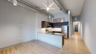 Located at 1630 Merchant Ln in Hampton, VA, this contemporary loft-style residence offers a thoughtfully designed single-floor layout with one bedroom, one bathroom, and a dedicated office. The space immediately impresses with its soaring ceilings, exposed industrial ductwork, and light hardwood flooring that run throughout, creating a warm yet modern aesthetic.

The open-concept kitchen and dining area serves as the heart of the home, featuring dark wood cabinetry, a classic white subway tile backsplash, granite countertops on the peninsula island with a double sink, and a full suite of stainless steel appliances. The adjacent living room benefits from the same generous ceiling height, with pendant lighting and a ceiling fan adding both function and style. A striking sliding barn door connects the living area to the rest of the unit, lending an industrial-chic character.

The bedroom is a well-proportioned retreat with high transom windows that allow natural light while maintaining privacy. A dedicated closet provides convenient storage. The office offers a versatile flex space with hardwood floors and neutral walls. The full bathroom is cleanly finished with a tub-shower combination, dark wood vanity, granite countertop, and light-toned flooring. Large glass entry doors at the front of the unit usher in abundant natural light and outdoor views.