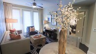 This single-floor living space showcases a thoughtfully designed interior that balances contemporary style with comfortable functionality. The living area serves as the focal point, featuring large windows dressed with white blinds and flowing curtains that allow abundant natural light to fill the room, creating a bright and airy atmosphere.

A ceiling fan with a built-in light fixture anchors the space from above, providing both practical airflow and a subtle design element. The room is furnished with a plush sofa and a classic armchair with an orange accent pillow, arranged around a distinctive rustic wood block coffee table that adds an organic, textural contrast to the otherwise modern aesthetic.

A bold black-and-white geometric area rug grounds the seating area, while a dark wood media console along the wall offers display and storage space. Decorative spherical accents and books add personality to the arrangement. A tall geometric faceted vase holding a dramatic white blossoming branch serves as a striking focal point, infusing the space with elegance. The soft gray walls and coordinated furnishings contribute to a cohesive, sophisticated ambiance throughout.