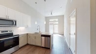 This single-floor residence is defined by its notably tall ceilings, which lend an airy, open quality throughout the living spaces. The open-concept kitchen is a central highlight, featuring an L-shaped layout with two-toned cabinetry — crisp white upper cabinets paired with warm greige lower cabinets — and granite countertops that extend along the peninsula. Pendant lighting hangs above the sink area, complementing the stainless steel appliances, which include a range with microwave, dishwasher, and refrigerator. A white subway tile backsplash adds a clean, classic finish.

The kitchen flows seamlessly into the living area, where rich wood-look flooring runs continuously underfoot. A set of French doors fitted with blinds serves as the primary entry point to the living space, topped with a transom window that draws in generous natural light. A ceiling fan overhead adds to the room's functionality.

The bathroom features a granite-topped vanity with undermount sink, a large framed mirror, and a full tub-shower combination with tiled surround. Wood-look flooring carries through here as well, maintaining a cohesive aesthetic. Overall, the space presents a clean, contemporary design with a bright, neutral palette throughout.