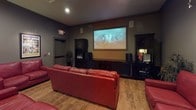 This single-level entertainment space is designed with a distinct cinematic theme, combining a home theater setup with a comfortable lounge and meeting area. The room is finished with dark gray walls and warm wood-tone flooring throughout, creating a moody, immersive atmosphere reminiscent of a private screening room.

The focal point of the space is a large wall-mounted projection screen flanked by a dark media console housing speakers and audio-visual equipment. Multiple red leather sofas, loveseats, and accent chairs are arranged in a tiered viewing configuration, offering generous seating capacity for group screenings or gatherings.

Classic Hollywood movie posters — including framed prints of Godzilla, King Kong, Scarface, and Rebel Without a Cause — are mounted throughout the room, reinforcing the cinematic identity of the space. Accent lighting, including table lamps and recessed ceiling lights, provides warm, dimmable illumination suited for both viewing and socializing.

A large dark wood conference or game table with chairs occupies one side of the room, offering a versatile area for meetings or activities. Exit signage and dedicated doorways connect the room to adjacent spaces, supporting its use as a shared or semi-public facility.