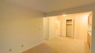 This single-floor space features a straightforward, functional layout with neutral tones throughout. The main living area is finished with wall-to-wall beige carpeting, smooth white walls, and standard-height ceilings, creating a clean and understated atmosphere. A partial wall divides the living space from an adjoining room, adding subtle architectural definition without fully closing off the floor plan.

The adjoining room, visible through the open passageway, appears to serve as a bedroom or sleeping area. It features a built-in closet with white bi-fold doors, offering organized storage with visible hanging rods and shelving. Adjacent to the closet, white cabinetry provides additional storage, suggesting a practical and efficient use of space. The carpeting continues seamlessly from the main area into this room, maintaining a consistent look throughout.

Overhead lighting in the secondary room provides warm illumination, contrasting with the cooler, ambient light of the main area. The overall design is minimal and utilitarian, with white trim and baseboards framing each room. The space is vacant, allowing for a clear view of its layout and structural features, making it easy to assess its potential for furnishing and personalization.