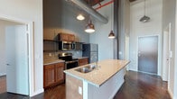 Located within the historic Germantown Mills Lofts, this single-floor loft unit showcases a compelling blend of industrial character and modern comfort. The open-concept kitchen serves as a central highlight, featuring warm cherry cabinetry, granite countertops, and a large island with a double-basin sink — ideal for both meal prep and casual gathering. Stainless steel appliances, including a range, microwave, and refrigerator, are complemented by open metal shelving that adds an industrial edge.

Soaring ceilings draw the eye upward to exposed ductwork, red-painted pipes, and original structural columns, all hallmarks of a thoughtfully preserved mill conversion. Dark hardwood floors ground the space and contrast beautifully with the cool gray walls. Vintage-style pendant lights with copper finishes hang over the kitchen area, providing warm, focused illumination while reinforcing the loft's industrial aesthetic. The generous ceiling height amplifies the sense of openness throughout, lending the space an airy, expansive feel characteristic of historic mill loft architecture.