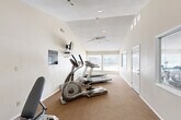 This fitness center at 715 Cedar Lake Rd SW in Decatur, Alabama, offers a dedicated exercise space designed to support a range of workout routines. The room features vaulted ceilings that lend a sense of openness, complemented by a ceiling fan that promotes air circulation during workouts. Neutral-toned walls and warm carpeted flooring throughout the main exercise area create a clean, comfortable environment, while a section near the entrance transitions to hard surface flooring.

Natural light fills the space through a generous bank of windows along one end, offering views of the surrounding community and keeping the atmosphere bright and inviting. A wall-mounted television provides entertainment or instructional viewing during cardio sessions.

The room is equipped with multiple treadmills and an elliptical trainer arranged along the carpeted area, ideal for cardiovascular exercise. A weight bench and a rack of dumbbells provide options for strength training, making this a versatile facility for a full-body workout. Posted exercise room use guidelines are displayed on the wall, ensuring users are informed of proper usage protocols. The space is accessed through a paneled entry door, and recessed and pendant lighting supplement the natural light for early morning or evening use.