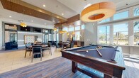 This single-story clubhouse in Sarasota, Florida, is a masterfully designed community gathering space that blends upscale architectural details with warm, resort-inspired interiors. The grand entrance hall immediately establishes the tone, showcasing soaring barrel-vaulted ceilings clad in rich wood, a dramatic starburst chandelier, and substantial stacked-stone columns flanking floor-to-ceiling glass walls. Natural travertine stone flooring flows throughout this central corridor, drawing the eye toward walls of windows that frame outdoor views and flood the space with natural light.

Adjacent to the entry, a reception area features a curved dark wood desk, teal accent walls, and elegant pendant lighting set against decorative shelving. Nearby, glass-walled conference rooms offer herringbone wood accent walls and professional furnishings for private meetings.

The expansive lounge and game room combines comfortable seating arrangements—including tufted and velvet upholstered chairs—with a full-size billiards table, wood-paneled ceilings, and large windows overlooking the surrounding landscape. A casual dining nook incorporates reclaimed wood walls, built-in banquette seating, and a striking geometric chandelier.

A fully appointed catering kitchen rounds out the amenities, featuring marble countertops, teal subway tile backsplash, wood cabinetry, and direct outdoor access—ideal for hosting events throughout this thoughtfully curated facility.