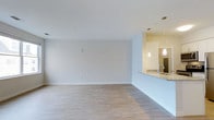 This studio features a single-floor open layout that maximizes usable space through a seamless flow between the living area and kitchen. The living space is finished with light hardwood-style flooring that runs throughout, complementing the crisp white walls and white ceiling to create a bright, airy atmosphere. Clean baseboards and a neutral color palette provide a versatile backdrop adaptable to a variety of interior styles.

The kitchen is positioned along one side of the space and is delineated by a raised counter that doubles as a breakfast bar, offering both functionality and a natural visual boundary between cooking and living areas. A granite or stone-look countertop adds a refined detail to the kitchen zone. Above the counter, two elegant pendant lights hang at staggered heights, providing focused task lighting while also serving as a design accent. A standard sink with modern fixtures is visible at the counter, and a mirrored or reflective surface nearby enhances the sense of depth and light within the kitchen area.

Overall, the space presents a clean, contemporary aesthetic well-suited for efficient, modern living.