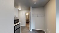 This single-floor unit offers a clean, modern design with a practical and efficient layout. The main living area features dark hardwood-style flooring that contrasts neatly with the light gray walls and white trim, creating a crisp, contemporary aesthetic. A built-in kitchenette provides everyday convenience, equipped with white upper cabinetry, a countertop, a microwave, and a compact refrigerator — all finished in a cohesive modern style. Adjacent to the kitchenette, an open closet area offers overhead wire shelving and a hanging rod, providing straightforward and accessible storage. Flush-mount ceiling light fixtures illuminate the space evenly, contributing to a clean and well-lit environment. A doorway leads directly into a private bathroom, which features wall-mounted vanity lighting above a sink, offering a functional and tidy layout. The bathroom's neutral tones maintain the consistent modern palette seen throughout the unit. Overall, this space is thoughtfully designed to maximize functionality within a compact footprint, making efficient use of every area while maintaining a cohesive, contemporary look.