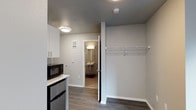 This single-floor unit offers a clean, modern design with a practical and efficient layout. The main living area features dark hardwood-style flooring that contrasts neatly with the light gray walls and white trim, creating a crisp, contemporary aesthetic. A built-in kitchenette provides everyday convenience, equipped with white upper cabinetry, a countertop, a microwave, and a compact refrigerator — all finished in a cohesive modern style. Adjacent to the kitchenette, an open closet area offers overhead wire shelving and a hanging rod, providing straightforward and accessible storage. Flush-mount ceiling light fixtures illuminate the space evenly, contributing to a clean and well-lit environment. A doorway leads directly into a private bathroom, which features wall-mounted vanity lighting above a sink, offering a functional and tidy layout. The bathroom's neutral tones maintain the consistent modern palette seen throughout the unit. Overall, this space is thoughtfully designed to maximize functionality within a compact footprint, making efficient use of every area while maintaining a cohesive, contemporary look.