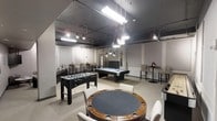 This single-floor entertainment space is thoughtfully designed to serve as a premier recreational hub. The open-plan layout accommodates a variety of gaming and social activities, anchored by a full-size pool table positioned at the center of the room beneath contemporary pendant lighting. A foosball table and a dedicated poker table with surrounding chairs provide additional entertainment options, catering to a range of interests and group sizes.

The industrial-style ceiling is left exposed, showcasing visible ductwork and pipework, while a combination of fluorescent strip lights and hanging pendant fixtures creates a well-lit yet atmospheric environment. The neutral color palette — featuring white and grey tones on the walls and light-toned flooring — keeps the space feeling clean and modern.

Toward the back of the room, a lounge area with comfortable seating and a flat-screen television offers a more relaxed setting for socializing or watching events. High-top tables with bar stools provide casual seating options near the perimeter. The space benefits from natural light filtering in through glass-paned doors at the rear, adding brightness and a sense of openness. Overall, the layout and furnishings make this an engaging, versatile space suited for community recreation and social events.