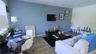 This single-floor space offers a thoughtfully designed interior that balances comfort and modern style. The living area serves as the focal point, finished with warm wood-look flooring that contrasts beautifully against cool blue-grey accent walls. A wall-mounted flat-screen television anchors the main entertainment area, complemented by a sleek dark media console styled with decorative vases and accents. A black lacquered coffee table sits atop a plush dark area rug, surrounded by white upholstered seating that adds a crisp, clean contrast to the room's palette. Framed artwork adorns the walls, lending a curated, personal touch to the space. A dedicated workspace is seamlessly integrated into the living area, featuring a white ergonomic chair and a compact desk, making the space as functional as it is visually appealing. Decorative elements such as dried floral arrangements and tasteful tabletop accessories add warmth and character throughout. The textured ceiling adds an architectural detail, while the overall layout provides a sense of openness and ease of movement. This space is well-suited for both relaxed everyday living and productive work, presenting a versatile and inviting environment.