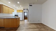 This single-floor unit offers a practical and functional layout across several distinct living areas. The kitchen serves as a central hub of the space, outfitted with warm honey-toned wood cabinetry extending to the upper walls, a full suite of white appliances including a refrigerator, electric range, and dishwasher, as well as a double-basin sink and a peninsula-style island that provides additional counter space. Recessed lighting illuminates the kitchen, which features a textured ceiling and durable light-toned laminate wood flooring that extends throughout the main living areas. The open layout connects the kitchen to an adjacent living or dining area, which is bright and neutrally finished with white walls and clean white baseboards. A separate carpeted room, featuring a bold geometric-patterned carpet in amber and brown tones, offers a warm and distinct contrast to the rest of the space and includes a wall-mounted television setup. The bathroom is equipped with a wood-toned vanity, a solid white countertop, an undermount sink with a brushed nickel faucet, and a large wall mirror. Throughout the unit, windows with horizontal blinds allow natural light to filter in, contributing to a bright and livable atmosphere.