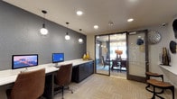 The business center and conference room is a thoughtfully designed, single-floor facility that blends functionality with contemporary style. The business center area features a long, built-in white countertop workstation lined with dark cabinetry below, providing ample workspace and storage. A desktop computer workstation is positioned along the desk, accompanied by a comfortable ergonomic chair, making it well-suited for tasks requiring focused productivity. Three stylish pendant lights with open cage-style shades hang overhead, casting warm, directed light over the work surface. The accent wall behind the workstation is finished in a deep charcoal textured treatment, adding visual depth and a modern character to the space. Patterned carpet tiles in neutral tones cover the floor throughout. Adjacent to the business center, a glass-walled conference room is accessible through a full-length sliding glass door, offering transparency while maintaining separation. The conference room features a round dining-style table surrounded by upholstered chairs, with natural light streaming in through a large window. A floral centerpiece adds a welcoming touch to this meeting area.