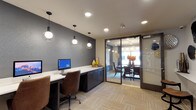The business center and conference room is a thoughtfully designed, single-floor facility that blends functionality with contemporary style. The business center area features a long, built-in white countertop workstation lined with dark cabinetry below, providing ample workspace and storage. A desktop computer workstation is positioned along the desk, accompanied by a comfortable ergonomic chair, making it well-suited for tasks requiring focused productivity. Three stylish pendant lights with open cage-style shades hang overhead, casting warm, directed light over the work surface. The accent wall behind the workstation is finished in a deep charcoal textured treatment, adding visual depth and a modern character to the space. Patterned carpet tiles in neutral tones cover the floor throughout. Adjacent to the business center, a glass-walled conference room is accessible through a full-length sliding glass door, offering transparency while maintaining separation. The conference room features a round dining-style table surrounded by upholstered chairs, with natural light streaming in through a large window. A floral centerpiece adds a welcoming touch to this meeting area.