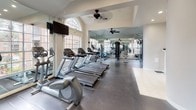 This fitness center is thoughtfully designed to accommodate a full range of workout needs within a bright, open layout. The space is divided into distinct functional zones, with a dedicated cardio area housing a row of treadmills, ellipticals, and stationary bikes, and a separate strength training section equipped with cable machines, weight benches, and free weights including a stability ball. The dark, polished concrete flooring in the main workout areas transitions to light ceramic tile near the entry, which features classic white French doors that allow natural light to pour in from outside. Large, multi-pane windows line the perimeter, offering views of the surrounding brick exterior and landscaping while flooding the interior with daylight. Vaulted ceilings with an arched transom window contribute to the airy, spacious feel of the room, and multiple ceiling fans maintain comfortable air circulation throughout. Floor-to-ceiling mirrors are mounted along the walls, visually expanding the space and allowing users to monitor their form during exercise. Wall-mounted televisions provide entertainment for cardio sessions. The overall ambiance is clean, bright, and functional, presenting a professional fitness environment suitable for a variety of training routines.