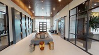 This contemporary amenity space makes a strong first impression with its single-floor layout that seamlessly integrates multiple functional areas through thoughtful architectural design. The main lobby entry features large-format light tile flooring, soaring ceilings clad in rich dark wood planks, and recessed lighting that creates a warm yet refined ambiance. Expansive floor-to-ceiling windows and dark-framed glass entry doors flood the space with natural light while offering open views of the surrounding exterior.

A statement feature wall dominates one side of the lobby, composed of large sculpted metallic panels with subtle backlighting, giving the space an artistic, high-end character. Curated lounge furnishings — including a low-profile gray sofa and unique organic wood and metallic accent stools — provide a comfortable gathering area.

The fully equipped fitness center is accessible directly from the lobby through steel-framed glass walls and doors, offering full visibility into the workout space, which features a comprehensive array of strength and cardio equipment. On the opposite side, a sliding barn-style door and glass partition wall lead to a separate clubroom, visible as a well-appointed social lounge. The cohesive use of black steel framing, warm wood tones, and neutral finishes throughout delivers a polished, modern aesthetic.