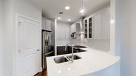 This single-floor space showcases a thoughtfully designed modern kitchen with a clean, contemporary aesthetic. The kitchen is anchored by a curved island topped with white quartz countertops featuring subtle speckled detailing, housing a double stainless steel undermount sink with a sleek arc faucet. Two decorative teardrop-style glass pendant lights hang above the island, adding an elegant visual focal point.

The cabinetry features a striking two-tone design, with light gray upper cabinets — some with glass-front panels for display storage — contrasting against dark charcoal lower cabinets. A subway tile backsplash runs along the walls, complementing the overall neutral color palette. The kitchen is well-equipped with stainless steel appliances, including a gas range, built-in microwave, and a French door refrigerator with an ice and water dispenser. A stainless steel wall-mounted range hood provides both function and a polished, professional look.

Recessed ceiling lighting brightens the space throughout, while rich dark hardwood flooring grounds the kitchen and flows into the adjacent open area. The light gray walls and white ceiling enhance the bright, airy ambiance of this well-appointed kitchen.