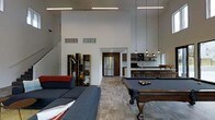 This single-floor space makes a striking impression with its soaring ceilings and open-concept layout, creating an expansive, airy atmosphere throughout. The main living area is anchored by a comfortable lounge with a large sectional sofa, seamlessly flowing into a recreation zone centered around a classic wood-framed billiards table — a standout feature perfect for entertaining guests.

Overhead, sleek linear pendant lighting and hanging Edison-style bulbs add both functional illumination and a modern industrial aesthetic to the space. The floors feature a light-toned wood-look finish that runs throughout, lending warmth and continuity to the open floor plan.

The kitchen is well-appointed with dark cabinetry, stainless steel appliances including a full-size refrigerator and microwave, open shelving, and a generous bar-style countertop with barstool seating — making it ideal for casual dining and socializing. A dedicated wet bar area adds further utility and entertainment value.

Subtle architectural details such as recessed ceiling vents and clean white walls keep the design minimal and modern. A staircase visible in the background hints at additional vertical space. Overall, this is a thoughtfully designed, multi-functional space well-suited for both relaxation and lively gatherings.