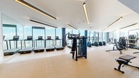 Located in the heart of downtown Miami at 700 NE 1st Ave, this high-rise fitness center offers a generously proportioned exercise space designed to accommodate a full spectrum of workout needs. The facility spans a single, open floor with a modern, contemporary aesthetic defined by clean white ceilings, light hardwood and low-pile carpeted flooring, and sleek linear pendant lighting throughout.

Floor-to-ceiling glass windows line the perimeter, flooding the space with abundant natural light and providing sweeping panoramic views of the Miami skyline, Biscayne Bay, and the surrounding cityscape — a standout feature that elevates the workout experience. Cylindrical structural columns in a deep navy finish add a bold architectural contrast to the otherwise bright and airy environment.

The gym is comprehensively equipped across several distinct zones. A dedicated cardio area features a long row of treadmills and elliptical machines positioned directly facing the city views. A separate cycling studio with light hardwood floors, full-length mirrors, and multiple stationary bikes offers a bright and open setting for indoor cycling. The strength training zone includes cable machines, a multi-station functional trainer, a full dumbbell rack, weight benches, and additional resistance machines. Large mirrors are strategically placed to expand the sense of space throughout.