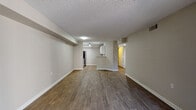 This single-floor unit is organized around a spacious, open-concept living and dining area finished with rich, dark hardwood-style flooring throughout. Neutral beige walls and white baseboards create a clean, cohesive aesthetic, while a textured popcorn ceiling runs consistently across the main living spaces. The layout flows naturally from the entry toward a peninsula-style kitchen, which features white shaker cabinetry, granite-patterned countertops, a stainless steel sink, and a stainless steel refrigerator. Track lighting overhead provides focused illumination in the kitchen area. A hallway off the main living space leads to additional rooms, including a carpeted bedroom that benefits from natural light through a full-width sliding glass door opening to a private patio or balcony with outdoor views. The bathroom is finished with matching hardwood-style flooring, a granite-topped vanity, and a full soaking tub with a white tile surround accented by a horizontal band of terracotta-toned tiles. The overall ambiance of the unit is functional and well-maintained, with consistent finishes throughout that give the space a modern, move-in-ready character.