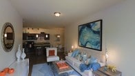 This single-floor residence features an open-concept layout that seamlessly integrates the living room, dining area, and kitchen into one cohesive and functional space. The living area is anchored by a comfortable neutral-toned sofa adorned with blue and patterned throw pillows, complemented by a plush accent chair in white with an orange cushion. A geometric-patterned area rug and a wood-topped coffee table add texture and warmth to the space. A large, bold abstract painting in deep blues and teals serves as a striking focal point on the main wall, contributing to the room's contemporary aesthetic. The kitchen features dark cabinetry, a breakfast bar with seating, and pendant lighting that adds an intimate ambiance. Track lighting further illuminates the workspace. Adjacent to the kitchen, the dining area is furnished with modern white chairs around a sleek table, bathed in warm light from a hanging pendant fixture. Flush-mount ceiling lighting throughout keeps the space well-lit, while the dark hardwood-style flooring provides a rich, grounding contrast to the lighter furnishings and walls. Overall, the space offers a modern, inviting atmosphere with thoughtful design details.