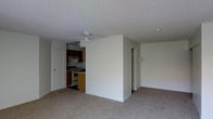 This single-floor unit offers a straightforward, functional layout designed for comfortable everyday living. The main living area is carpeted throughout, providing a warm underfoot feel, and is centered beneath a ceiling fan with integrated lighting — ideal for year-round comfort. The textured ceiling adds a classic touch to the space.

The kitchen is positioned adjacent to the living area and is partially separated by a half-wall partition, creating a semi-open feel while maintaining a sense of definition between the spaces. It features warm wood-toned cabinetry on both the upper and lower sections, a stainless sink set into a light-colored countertop, and a freestanding range with an overhead ventilation hood. The darker flooring in the kitchen contrasts with the carpeted living space, helping to visually distinguish the two zones.

A closet with a hanging rod is visible toward the rear of the living area, offering convenient built-in storage. Multiple doorways lead to additional rooms, suggesting a practical floor plan with separated private and common areas. The neutral white walls throughout keep the space feeling open and bright, making it easy to personalize. Overall, the unit presents a clean, no-frills layout well-suited for efficient, day-to-day living.