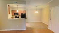 This single-floor residence offers a thoughtfully designed open-concept layout that connects the kitchen and living areas in a cohesive, functional flow. The kitchen is equipped with black appliances, including a refrigerator, range, and built-in microwave, complemented by warm wood cabinetry and a granite-topped breakfast bar that serves as both a prep space and a casual dining spot. A single pendant light hangs above the bar, providing focused task lighting with a modern touch. The transition from the kitchen into the living and dining areas is marked by a shift from hardwood flooring to plush wall-to-wall carpeting, adding warmth and comfort underfoot. The dining area is anchored by an elegant multi-bulb chandelier that adds a refined focal point to the space. Neutral, warm-toned walls throughout create a welcoming and versatile backdrop, allowing for a wide range of décor styles. Recessed ventilation and clean white trim details contribute to the polished, move-in-ready feel of the home. The overall ambiance is bright, open, and inviting, making it an ideal space for both everyday living and entertaining.