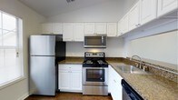 This single-floor space offers a thoughtfully designed kitchen that serves as a central feature of the home. The kitchen is outfitted with a full suite of stainless steel appliances, including a refrigerator, electric range with oven, over-the-range microwave, and dishwasher, providing everything needed for everyday cooking and meal preparation. Crisp white cabinetry lines the walls both above and below the countertops, offering generous storage space and contributing to a bright, clean aesthetic. The dark granite-style countertops provide a striking contrast against the white cabinets, adding a touch of warmth and visual depth to the space. A double-basin sink is conveniently positioned near the corner, complementing the efficient U-shaped layout that maximizes workspace and ease of movement. Vaulted ceilings in the kitchen area enhance the sense of openness and airiness, making the space feel larger than its footprint might suggest. The wood-style flooring grounds the room with a warm, natural tone that ties the overall design together cohesively. The overall ambiance is clean, functional, and inviting, reflecting a practical yet polished interior aesthetic well-suited for comfortable everyday living.