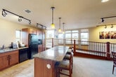 This thoughtfully designed multi-use suite offers a comfortable and functional layout across a single floor, seamlessly blending kitchen, dining, and lounge areas into one cohesive space. The kitchen is a standout feature, showcasing rich warm-toned cabinetry, granite countertops, and a large central island with bar seating illuminated by pendant lighting. Black appliances, including a full-size refrigerator, microwave, and dishwasher, are complemented by a sink with a granite surround and ample upper and lower cabinet storage. Track lighting adds a modern touch overhead. Adjacent to the kitchen, a spacious open lounge area features plush seating, a wall-mounted flat-screen television, and recessed lighting, creating a relaxed and inviting atmosphere. Large windows and sliding glass doors allow generous natural light to fill the room while offering views of the surrounding outdoor environment and access to an exterior balcony. A distinctive wrought-iron and wood staircase railing defines a loft-style upper overlook area, providing additional seating and display space accented by bold wall art and directional track lighting. The space also includes a formal dining table with multiple chairs, ideal for gatherings. Neutral carpet and tile flooring tie the various zones together for a warm, welcoming ambiance throughout.