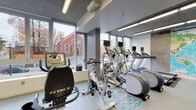 Located at 1726 SW 4th Ave in Portland, Oregon, this fitness center offers a comprehensive workout facility designed with both functionality and aesthetics in mind. The space features an exposed concrete ceiling with linear fluorescent lighting, lending it a modern industrial character throughout.

The cardio zone is positioned along a wall of large floor-to-ceiling windows, flooding the area with natural light and providing open views of the surrounding Portland streetscape, including a notable historic red brick building nearby. This section is equipped with treadmills, elliptical trainers, and stationary bikes, many fitted with individual media screens. A large decorative world map mural serves as a striking visual focal point along one wall.

The strength and free weights area is equally well-appointed, featuring adjustable weight benches, a full rack of dumbbells, medicine balls, resistance equipment, and a cable/pulley machine. Accent walls in teal and gray add visual interest to this section, which also benefits from continued natural light through additional windows.

The light wood-toned flooring, gray walls, and teal accents create a clean, energizing atmosphere throughout the facility, making it a well-rounded space for a full range of fitness activities.