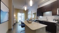 This single-floor residence showcases a thoughtfully designed open-concept layout that seamlessly connects the kitchen, dining, and living areas into one cohesive living space. The kitchen is a standout feature, boasting dark espresso cabinetry, a classic white subway tile backsplash, and a generous curved quartz-topped island with bar seating — perfect for casual dining or entertaining. Pendant lighting above the island adds a refined touch, complementing the track lighting that runs along the ceiling to illuminate the entire space. The living area flows naturally from the kitchen, furnished with comfortable seating and tasteful décor, creating a warm and inviting atmosphere. Large sliding glass doors at the far end of the space flood the interior with natural light and provide direct access to an outdoor area, framed by sheer yellow curtains that add a soft, warm accent. The flooring transitions between tile and wood-look surfaces, subtly defining each zone within the open plan. High ceilings throughout enhance the sense of spaciousness, while the neutral color palette with modern finishes gives the space a clean, contemporary feel. Overall, this residence offers a well-balanced blend of functionality and modern style.