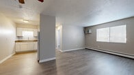 This single-floor unit offers a practical and straightforward layout designed for comfortable everyday living. The open living and dining area is finished with wood-look laminate flooring in a warm gray tone, complemented by neutral-painted walls and a textured ceiling. A ceiling fan provides overhead circulation, while a wall-mounted air conditioning unit and a baseboard heater ensure year-round climate control. A window allows natural light to fill the space, contributing to an airy feel. A structural half-wall separates the living area from the kitchen, which features white cabinetry, dark granite-style countertops, a double-basin sink, a white electric range, and a full-size refrigerator. Under-cabinet lighting adds a practical touch. A hallway, lit by a flush ceiling fixture, connects the living spaces to the private areas of the unit. The bedroom is a well-proportioned room with a sliding-door closet, a ceiling fan, and continued wood-look flooring. The bathroom is clean and functional, offering a full soaking tub and shower combination with white surround panels. Throughout, white trim and baseboards provide a cohesive, tidy finish.